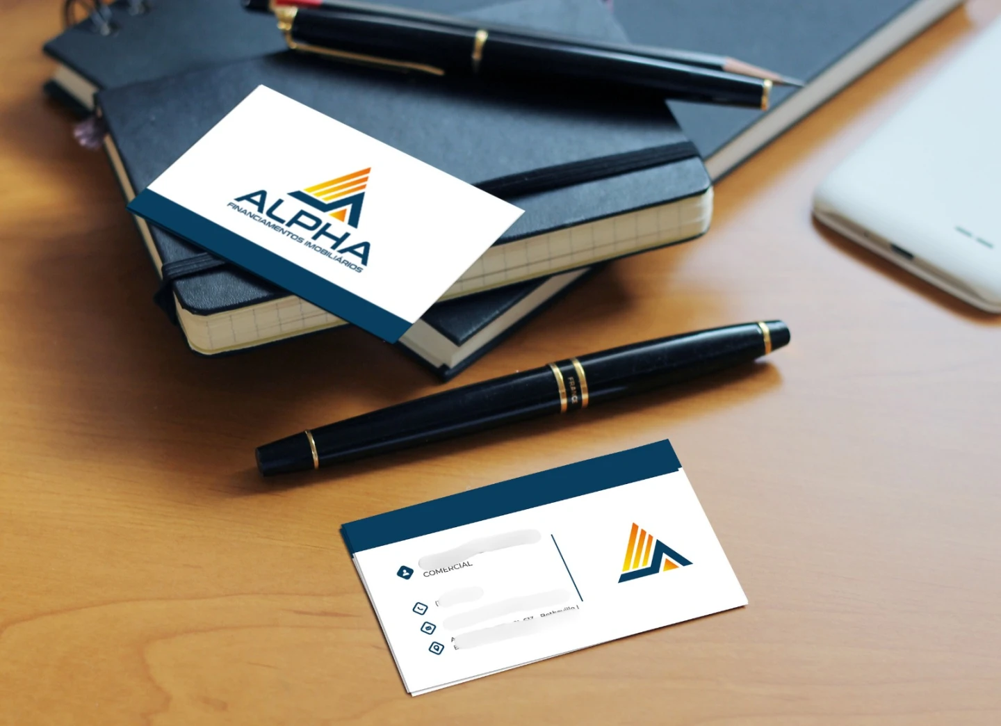 Cartões de visita institucionais impressos da Alpha Financiamentos Imobiliários dispostos sobre uma mesa de madeira ao lado de cadernos e canetas.