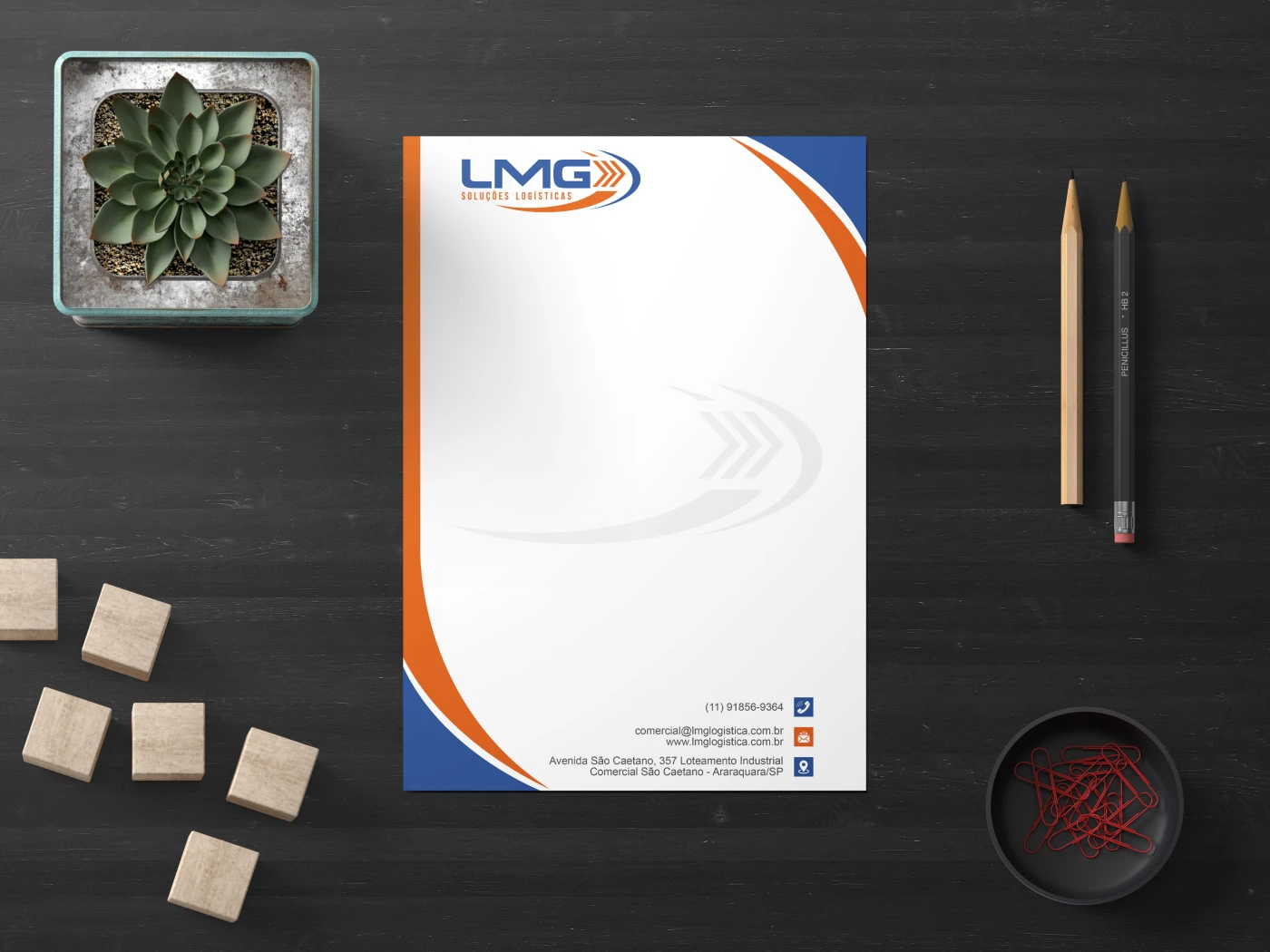 Papel timbrado da LMG Logística sobre mesa de escritório escura com lápis e acessórios, exibindo design em azul e laranja.